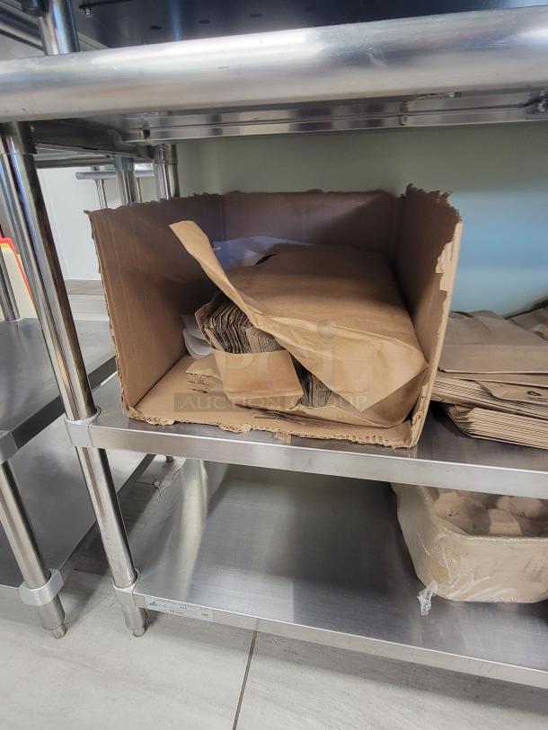 KCS WS-2460 stainless steel prep table with bulk brown paper bags, drink carriers, and plastic cutlery on lower shelf.