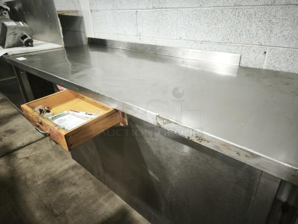 Stainless steel prep table with wooden drawer, in good condition. Features a clean surface and sturdy construction.