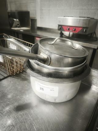 Several stainless-steel saucepans stacked, showing handles and lids. Visible tag labeled "26 QTY." Used condition.