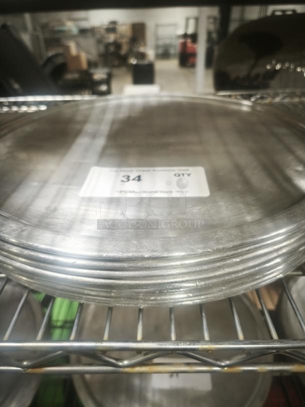 Stack of aluminum pizza pans on a metal shelf, showing light wear. Auction label reads "34 QTY, PCIAuctions.com."