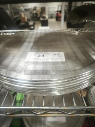 Stack of aluminum pizza pans on a metal shelf, showing light wear. Auction label reads "34 QTY, PCIAuctions.com."