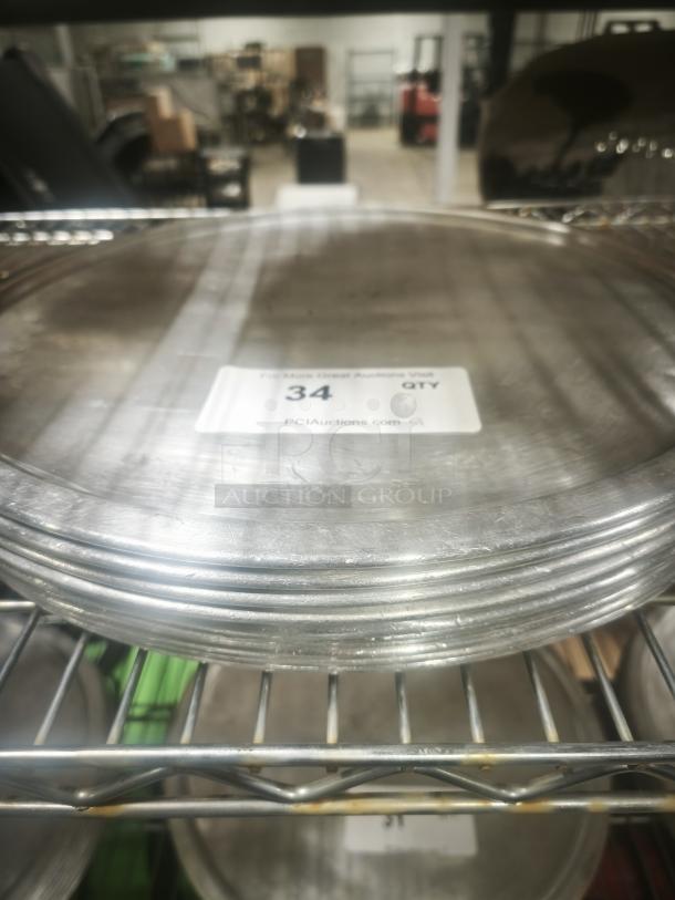 Stack of aluminum pizza pans, used condition, on metal rack. Label with "34 QTY PCIAuctions.com" visible.