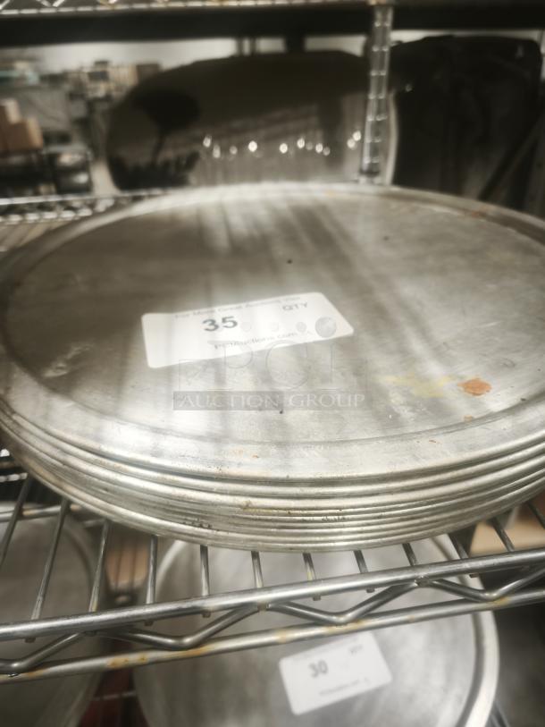 Stack of aluminum pizza pans, good condition, slight wear. Tags show quantity of 35. Stored on metal rack.