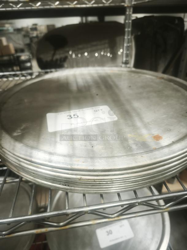 Stack of aluminum pizza pans on a metal shelf, with visible surface scratches and labels indicating quantity.