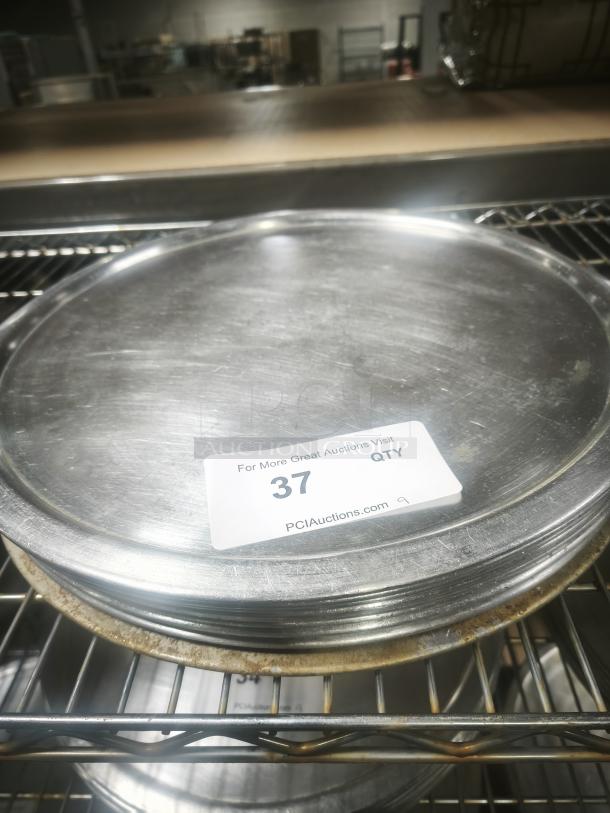 Stack of aluminum pizza pans on a metal rack, moderate wear, marked "PCI Auctions, 37 QTY," auction label visible.