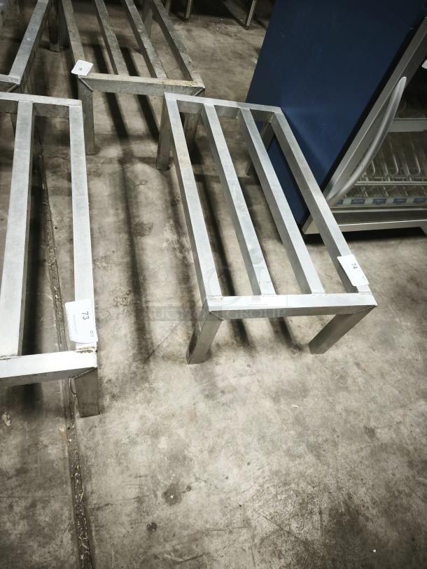 A collection of metal frames on a concrete floor, possibly used as industrial shelving or supports. Each frame has a visible auction tag with numbers.