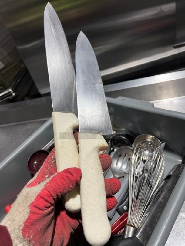 Lot of miscellaneous kitchen utensils, including two chef knives with white handles in good condition, whisks, and ladles, all in a grey bin.