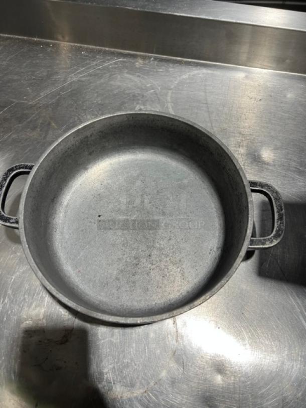 Commercial round braiser pan with handles and dome-style lid, shown in used condition on a metal surface.