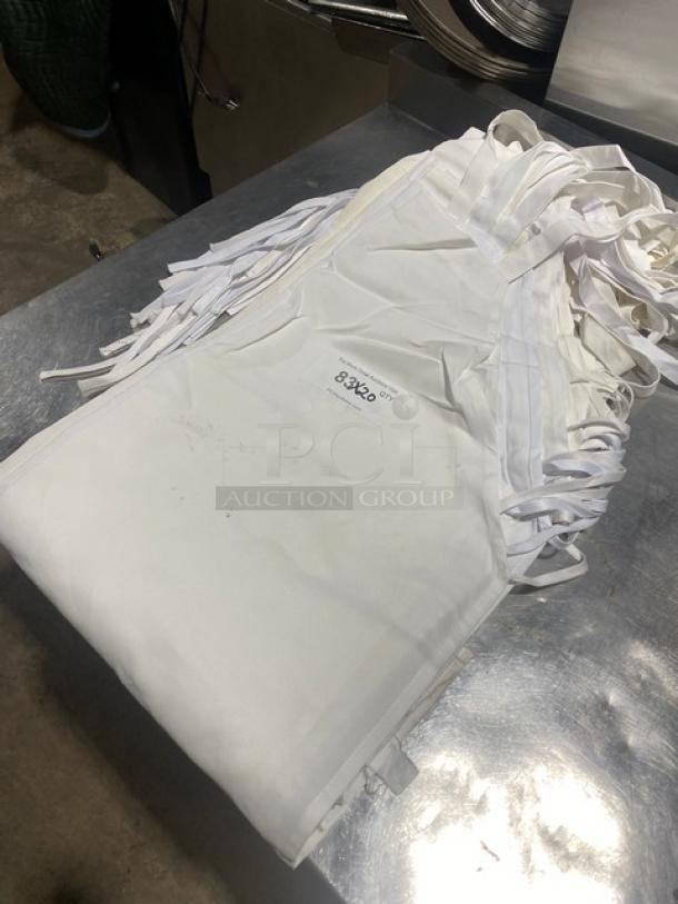 Stack of white full-length bib style aprons, new condition, labeled 33x20, commercial use, on metal surface.