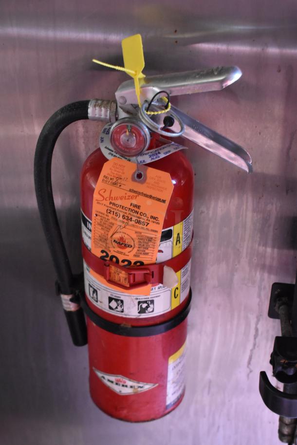 Red fire extinguisher with inspection tags and safety seal, mounted on metal wall inside trailer. Label: "Schweizer Fire Protection Co."
