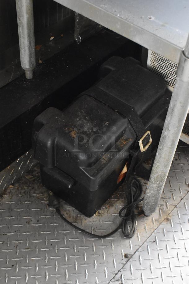 Black equipment case secured under a metal shelf inside the ATX 28' cold food and beverage trailer. Dirt visible on the case.