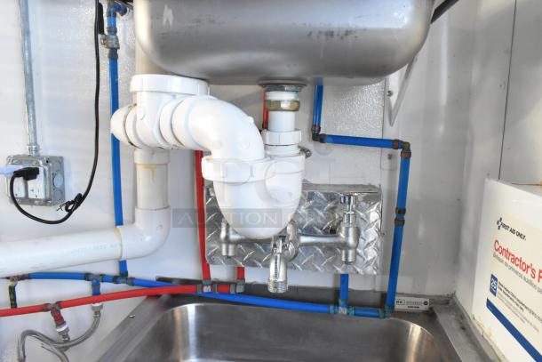 Interior view of retail trailer plumbing with stainless steel sink, white PVC piping, and electrical outlet. Contractor's supply box visible.