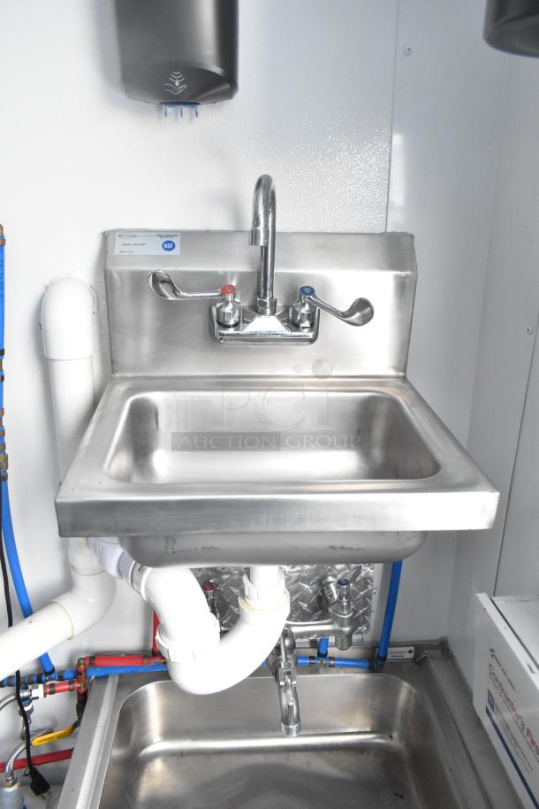 Stainless steel sink with dual-handle faucet in a retail trailer. NSF sticker visible; plumbing and fixtures shown.
