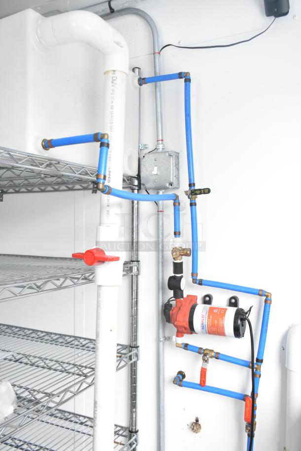 Interior of a cold food and beverage trailer, highlighting plumbing and electrical setup with metal shelving and red valve.