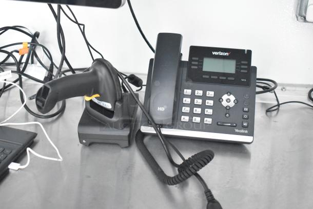 Yealink T41S desk phone with Verizon branding and HD label, placed on a metal surface. Near it is a barcode scanner on a dock, surrounded by various cables.