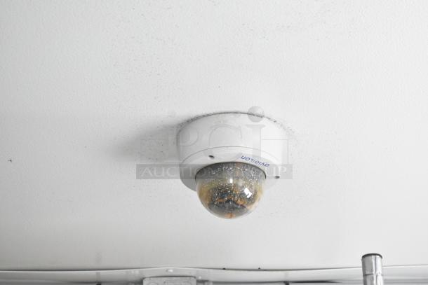 Ceiling-mounted surveillance dome camera in a trailer, branding partially visible, suggesting security features.