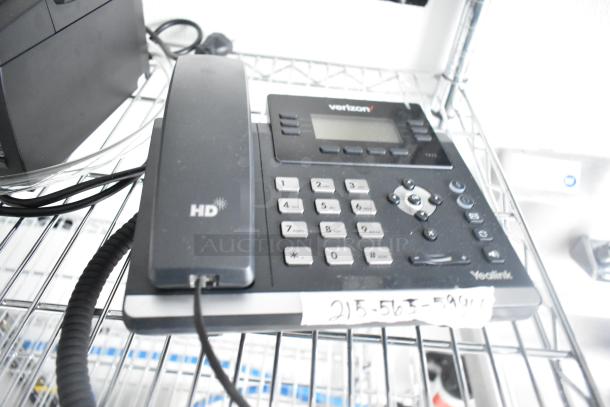 A Yealink HD office phone with a Verizon logo, displayed on a metal shelf, accompanied by a coiled handset cable.