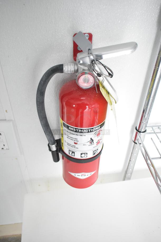 Red fire extinguisher mounted on a wall with instruction label and tags. Part of Nationwide 24' Shell Food Trailer equipment.