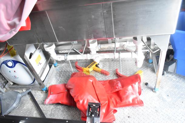 Alt text: "Interior view of a Titanium Cargo 28' food trailer with metal sink, plumbing, water heater, and red protective gear on diamond plate flooring."