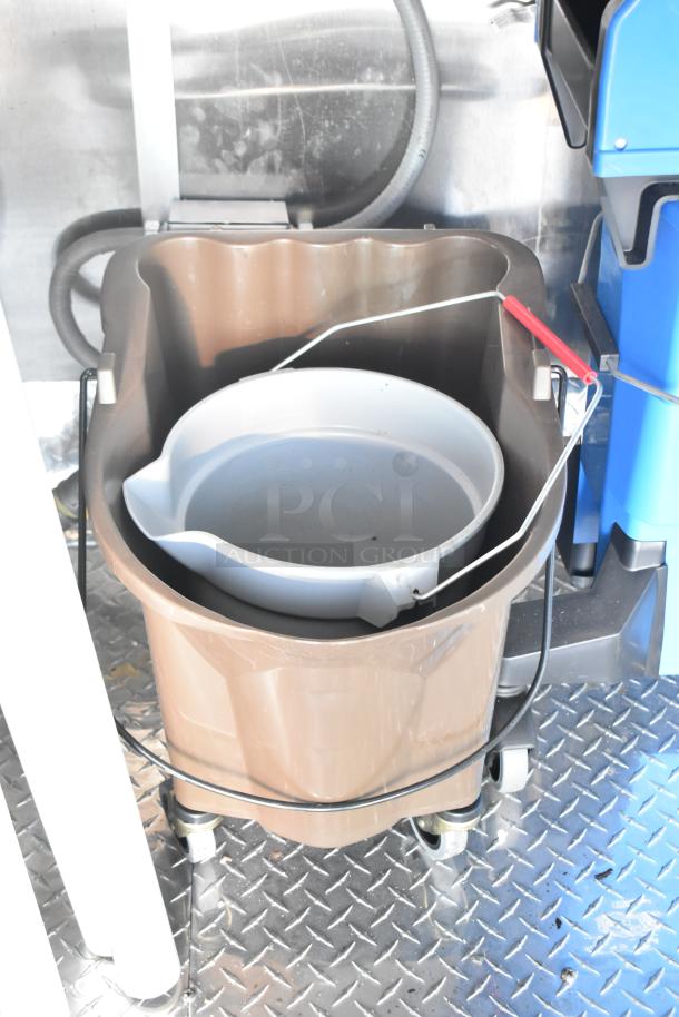 Brown mop bucket with a white wringer on metal casters inside a mobile kitchen trailer.