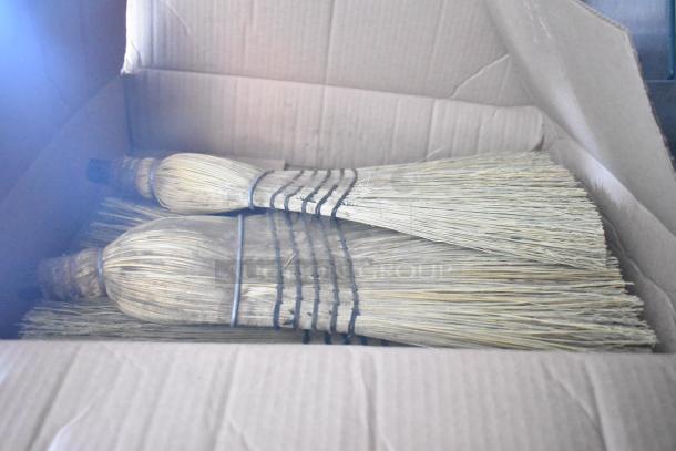 Cardboard box containing bundled straw brooms, secured with string ties, showcasing natural bristle texture.