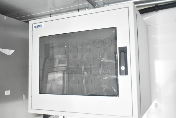 White metal cabinet with glass door and handle, brand "martin" visible on top corner. Inside are wire shelving units with various equipment.