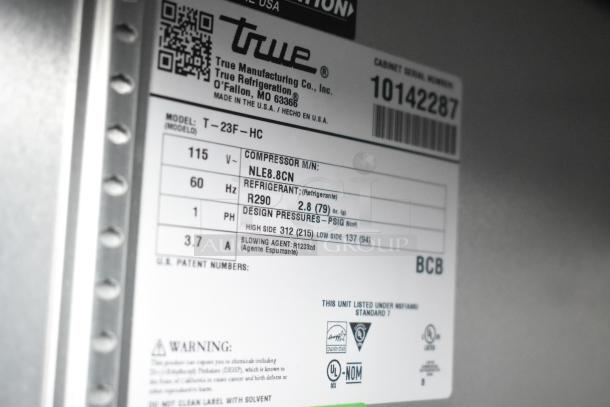 Close-up of a True model T-23F-HC refrigeration unit label, showing specifications like voltage, refrigerant type, and safety warnings.