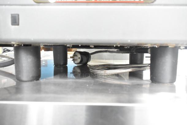 La Cimbali M29 Select DT/2-NF espresso machine underside, showing 208/240V power cord and legs on stainless steel surface.