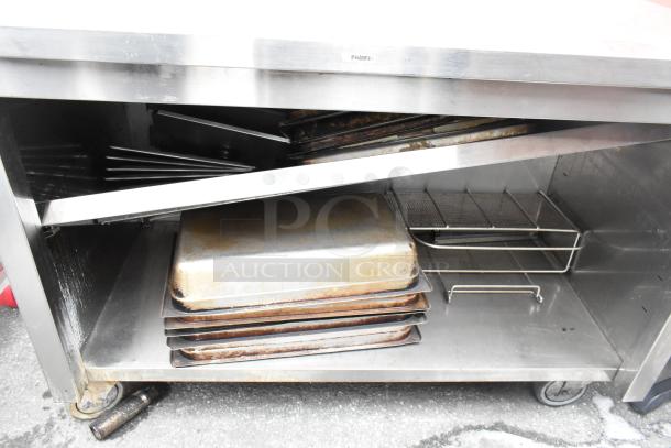 Stainless steel commercial table with backsplash, under shelf, and contents including trays and racks. Marked "Fridm" on edge.