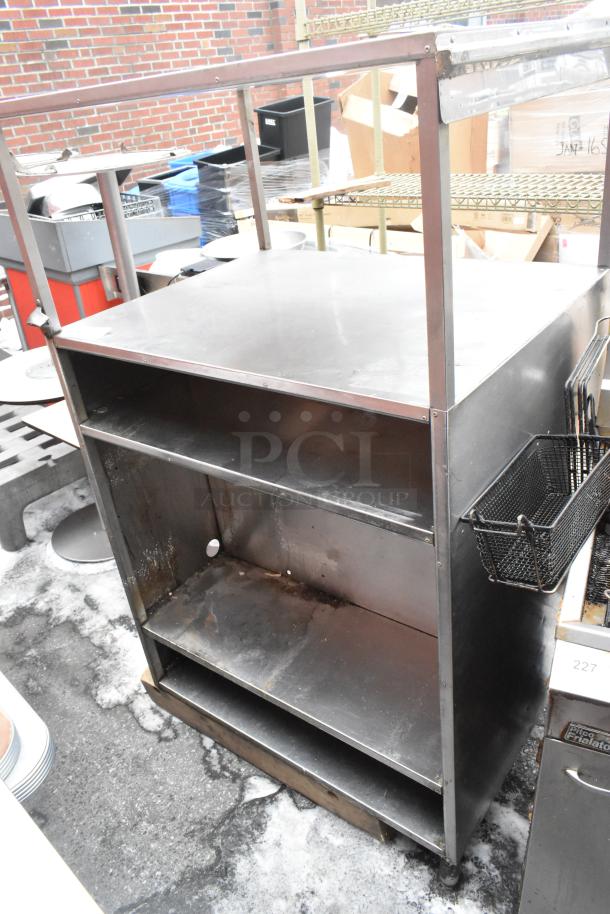 Stainless steel shelving unit with two shelves, surface wear visible, black wire basket attached to side.