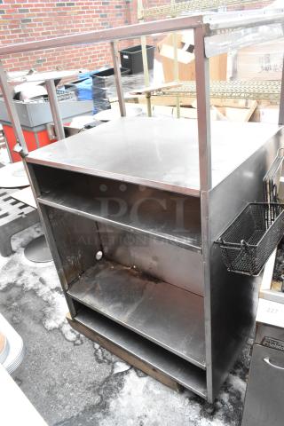 Stainless steel shelving unit with two shelves, surface wear visible, black wire basket attached to side.
