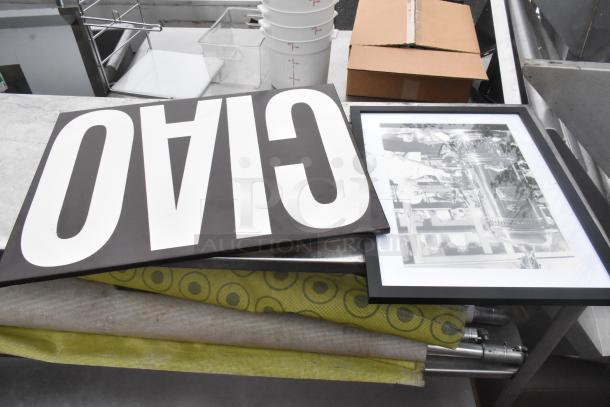 4-tier shelving unit with various contents including a framed black-and-white photo, a large black-and-white sign, and rolled textiles.