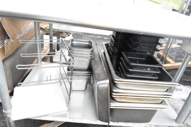 Commercial table with under shelf, holding various metal and plastic containers, sturdy metal frame, used condition.