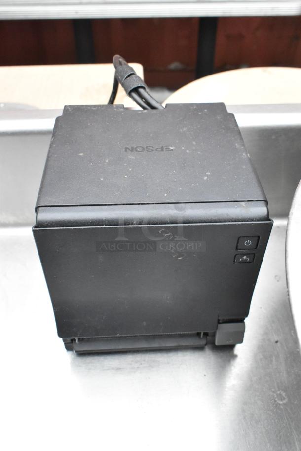 A black Epson device, likely a printer or similar, placed on a commercial table with visible power and paper feed buttons.
