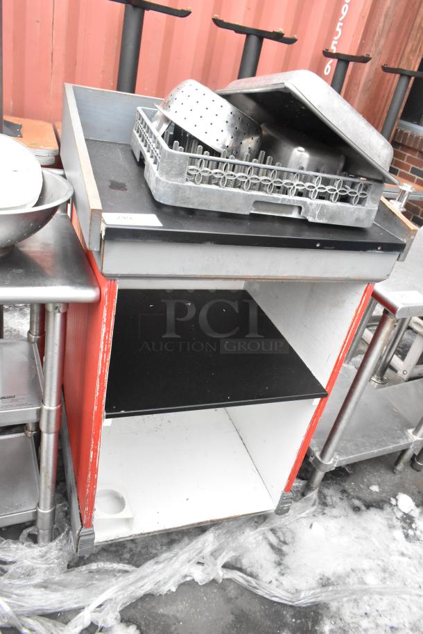 Metal counter with black top, red sides, and shelves containing kitchen trays and utensils. Visible wear on surfaces.
