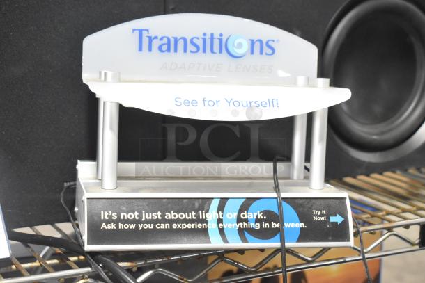 Display stand for Transitions Adaptive Lenses, featuring promotional text about light adjustment. Includes signage and cable connections.