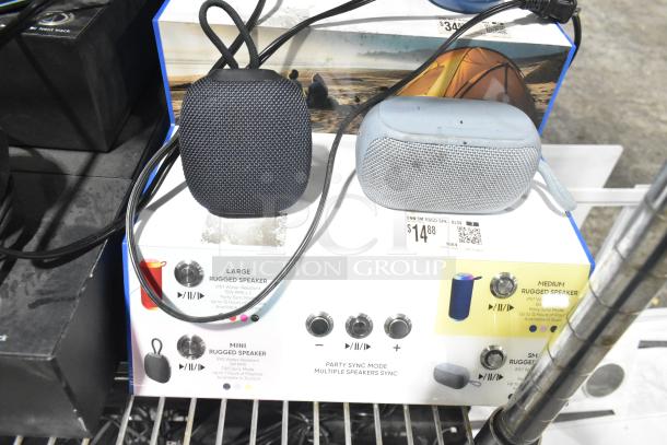 Two rugged speakers, one black and one gray, on a display with labeled features; Onn speaker priced at $14.88.