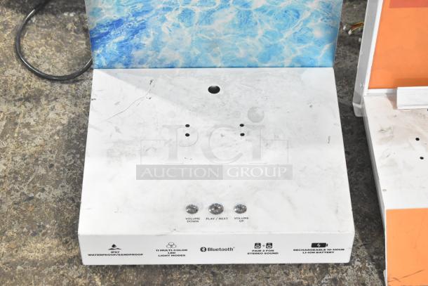 White display stand with blue background, featuring buttons labeled for volume and playback. Displays icons for Bluetooth, waterproof, and light modes.