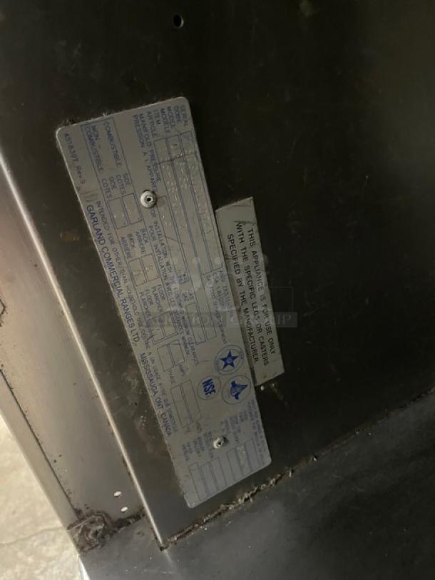 U.S. Range commercial stainless steel gas-powered 6-burner range with backsplash, storage base, on casters; visible tags with specifications.