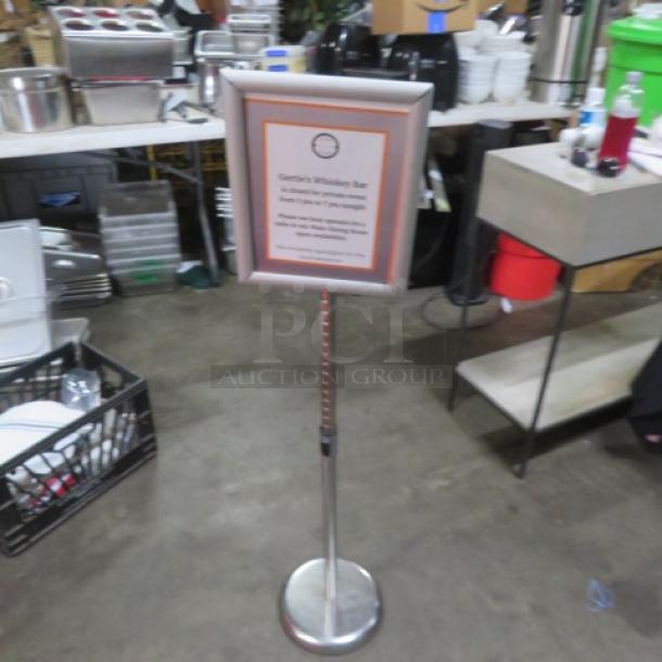Adjustable sign with silver frame on a pedestal base, holding a printed menu. Used, in good condition. Suitable for displays.