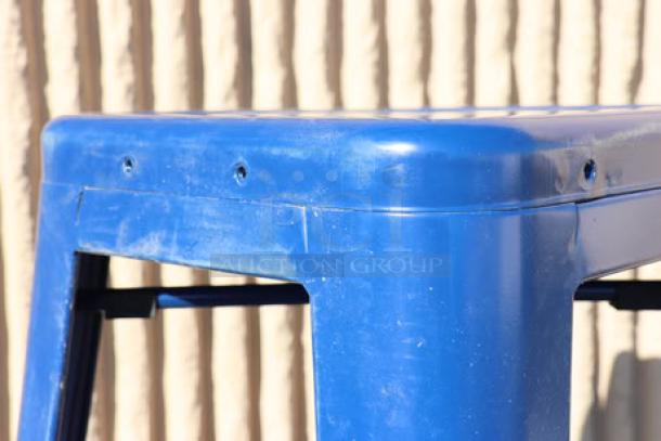 Blue metal stool with a backrest visible. Shows small dents and scratches, highlighting use. Set against a textured background.