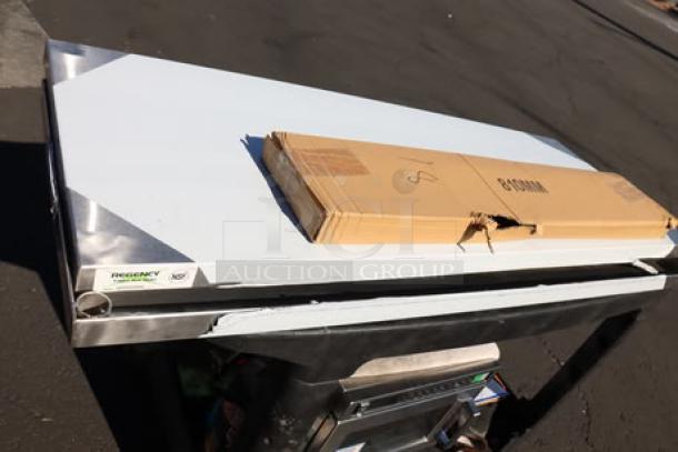 Regency stainless steel overshelf with scratches, visible "Regency" label. Brown cardboard on top. Appears unused.