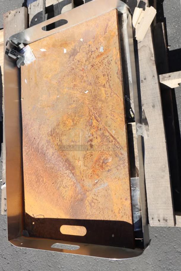 Steel griddle top with visible rust and scratches, placed on wooden pallet. Edges reinforced with metal trim, features two cut-out handles.