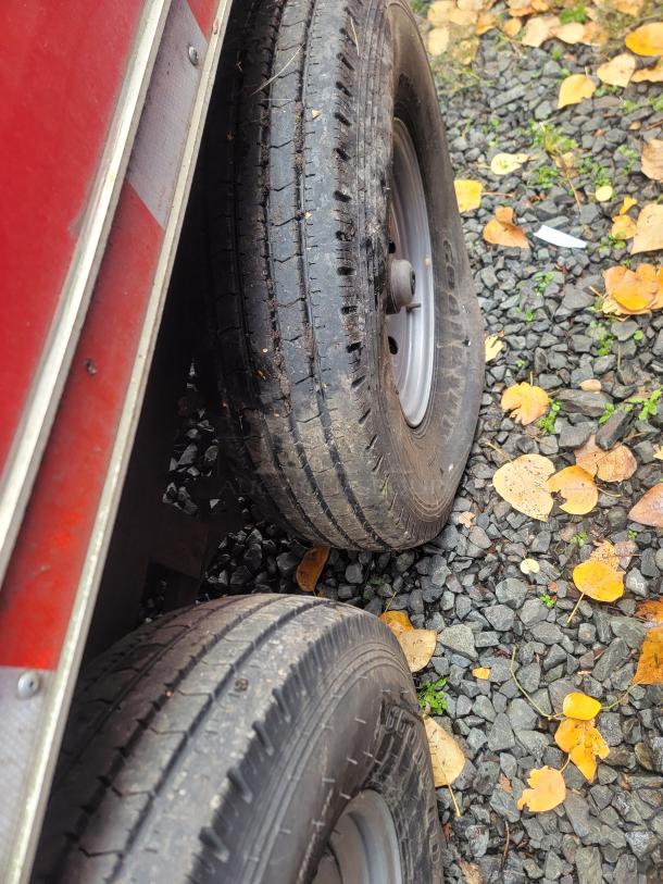 Red 2021 Titanium Cargo Trailer's double axle with black tires, surrounded by fallen autumn leaves on gravel.
