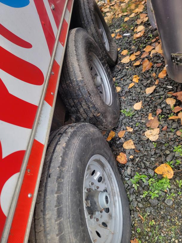 Triple axle wheels of the 2021 Quality 28' Wendy's Mobile Ghost Kitchen trailer on gravel with autumn leaves.
