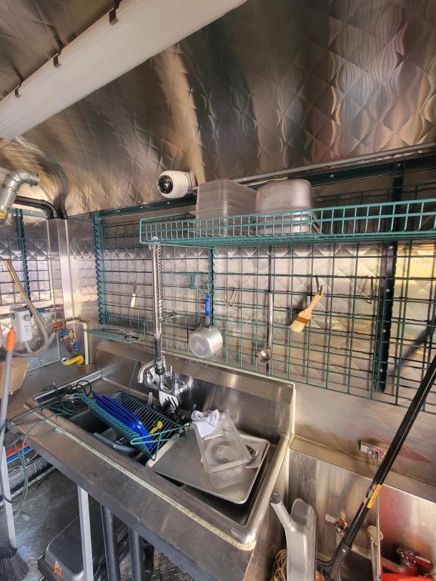 2021 Quality 24' food trailer kitchen interior with stainless steel build, three-compartment sink, surveillance camera, and storage rack.