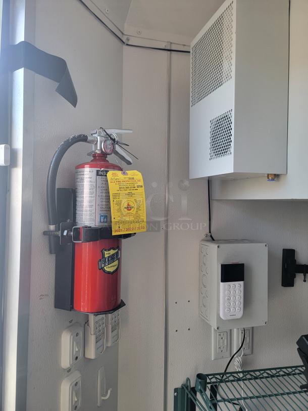 Fire extinguisher with safety inspection tag inside a mobile retail/freezer trailer, next to a keypad security system.