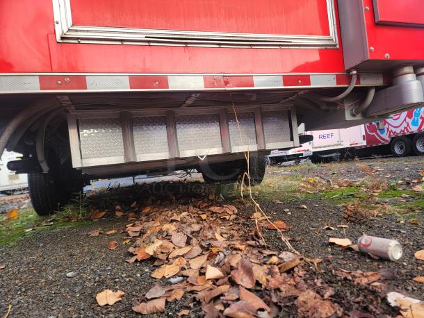 Underneath view of a red 2022 Rock Solid Cargo 28' triple-axle ghost kitchen trailer. Features include a diamond plate toolbox. The ground is covered with fallen leaves.