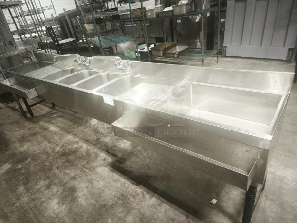 Stainless steel 4-bay bar sink in premium condition, featuring dual faucets and integrated drainboards. Ideal for commercial use.