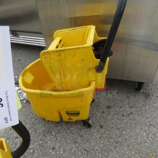 Yellow mop bucket with wringer, one missing caster, visible cleaning-related label, used condition.